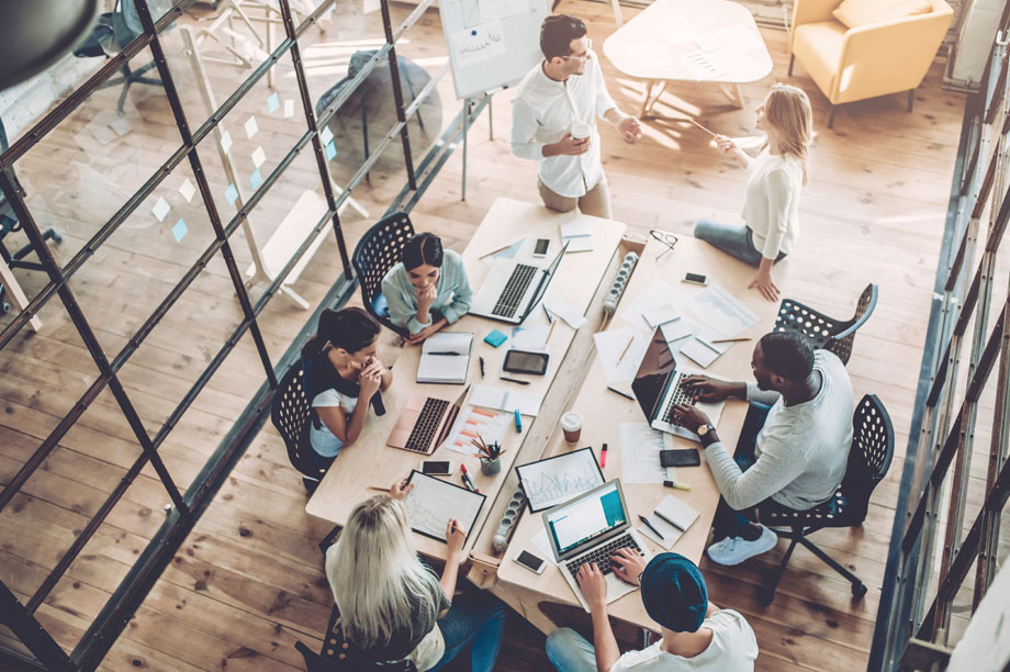 Image looking down on a group of learners around a large office desk with laptops collaborating on a project